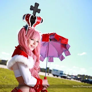 Kasia Northeast as Perona from One Piece, smiling in a pink wig and crown while holding a red parasol with a purple underside beside a grassy slope.