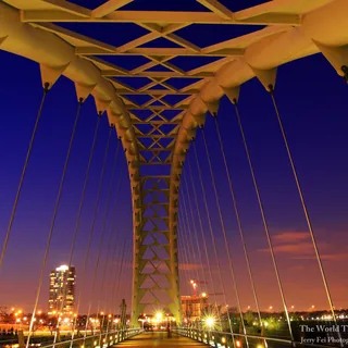 Golden-lit steel arches and crossing beams frame the pedestrian walkway of Humber Bay Arch Bridge, with suspension cables and Toronto waterfront lights beneath a deep blue evening sky.