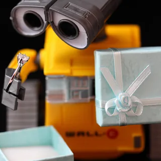 Close-up of a yellow WALL-E toy holding a clear-stone ring and a pale blue gift-box lid tied with white ribbon against a black background.