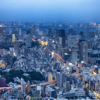 Illuminated Tokyo Tower above dense city blocks and curving expressways at blue hour, with Rainbow Bridge and Tokyo Bay visible in the distance.