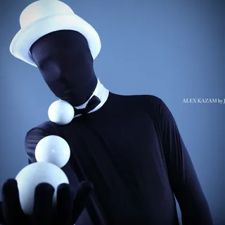 Alex Kazam in a dark face covering, white top hat, and black bow tie, balancing three white balls from his outstretched gloved hand along his arm to his shoulder.