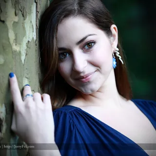 Ashley in a deep blue dress looking at the camera beside a textured tree trunk, with one hand resting on the bark, blue-painted nails, and a blue flower earring.