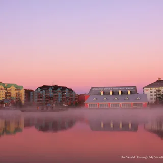 Muskoka Boat & Heritage Centre and neighbouring waterfront buildings reflected in misty Lake Muskoka beneath a pink-and-lavender sky at Muskoka Wharf in Gravenhurst.