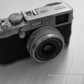 Silver-and-black Fujifilm X100S camera viewed from above at an angle on a pale wooden surface, showing its lens and top control dials in black and white.