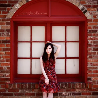 Woman in a red patterned dress and black heels posing before an arched, red-framed window in a brick wall, with one arm over her head.