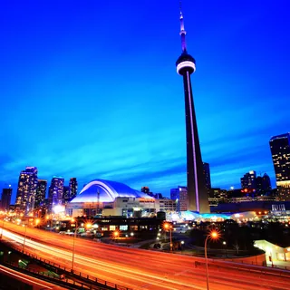 CN Tower and the domed Rogers Centre beneath a vivid blue twilight sky, with orange and white traffic light trails along Toronto’s Gardiner Expressway.