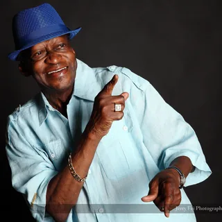 Smiling man wearing a blue fedora and pale blue button-up shirt, with one index finger raised and his other hand pointing downward against a dark background.