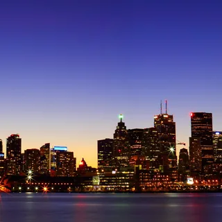 Wide panoramic Toronto skyline with the red-lit CN Tower across Lake Ontario, beneath a blue sky and orange horizon, with illuminated buildings and waterfront reflections.