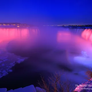 Horseshoe Falls illuminated in pink and violet beneath a deep blue sky, with drifting mist and snow and ice along the Niagara River in winter.