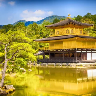 The golden upper floors of Kinkaku-ji reflected in Kyoko-chi Pond, with pine trees in the foreground and wooded hills beneath a blue sky in Kyoto.