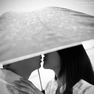 Grace and Ben leaning toward a kiss beneath a translucent umbrella in a black-and-white engagement portrait.