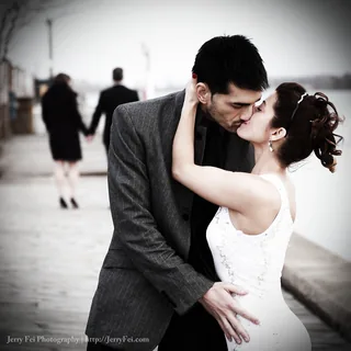 Couple in a gray suit and white dress kissing on a waterfront promenade, with another couple walking hand in hand behind them.