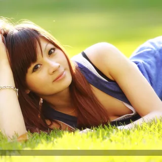 Jaymee Chen (陳怡慈) reclining on sunlit grass in a blue sleeveless top, with one hand resting in her hair and warm backlighting around her head.
