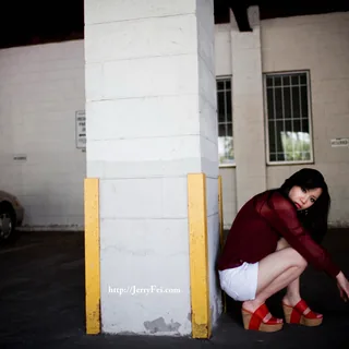 Woman in a burgundy blouse, white shorts, and red platform sandals crouching beside a concrete pillar with yellow protective strips in a covered parking area.