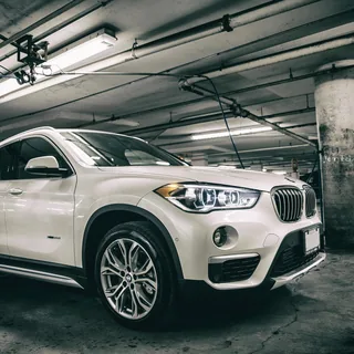 White BMW X1 photographed from a low front three-quarter angle in an underground parking garage, with illuminated headlights, concrete pillars, and overhead lights.