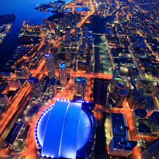 Aerial view of Toronto at dusk, with the blue-lit Rogers Centre in the foreground, glowing roads through CityPlace, and Billy Bishop Toronto City Airport beside Lake Ontario.