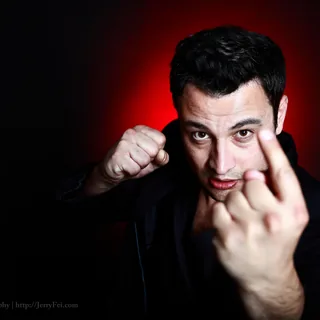 Man in a black jacket looking directly at the camera, with one fist raised and his other hand beckoning toward the lens, against a dark background with red light.