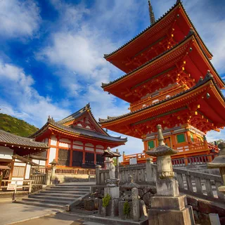 Vermilion three-story pagoda at Kiyomizu-dera in Kyoto, with ornate painted beams, neighboring temple roofs, stone steps, and stone lanterns beneath a blue sky.