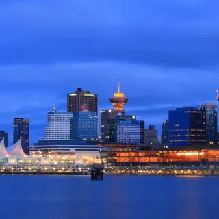 Vancouver waterfront beneath a blue evening sky, with the white sails of Canada Place, the illuminated Vancouver Lookout at Harbour Centre, and high-rise buildings above the water.